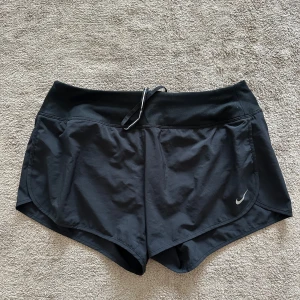 Svarta träningsshorts från Nike - Svarta träningsshorts från Nike med Dri-FIT-teknologi. Shortsen har en elastisk midja med snörning och liten dragkedjeficka bak. Perfekta för löpning eller gymmet.