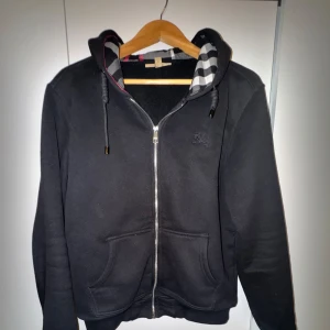 Svart hoodie med dragkedja från Burberry - Säljer en svart hoodie från Burberry med dragkedja och klassiskt rutigt foder i huvan. Tröjan har två fickor framtill och justerbar huva med snören. Perfekt för en stilren och bekväm look.