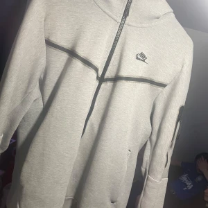 Ljusgrå zip-hoodie från Nike - Säljer en ljusgrå hoodie från Nike med hel dragkedja och svarta detaljer. Jackan har lång ärm, fickor framtill och en liten Nike-logga på bröstet. Perfekt för en sportig och avslappnad stil.