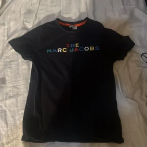 Svart t-shirt från Marc Jacobs - Svart t-shirt från Marc Jacobs med färgglad logotyptext på bröstet. Klassisk rund halsringning och korta ärmar. Perfekt för en stilren och trendig look.
