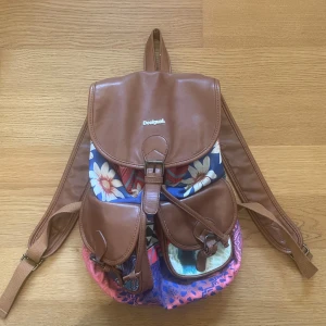 Desigual väska - Brun Desigual ryggsäck med färgglada detaljer och mönster. Inuti finns ett mönstrat foder med en liten ficka med dragkedja. Ryggsäcken har justerbara axelband. Köpt i Spanien för ca 9 år sedan och använd max 5 gånger. Ryggsäcken är nästan helt felfri förutom att färgen på öglan har försvunnit lite.