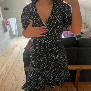 Svart kortklänning med vita prickar - Kort omlottklänning i svart med vita prickar och v-ringning. Klänningen har korta puffärmar och volang nertill, vilket ger en söt och trendig look. Perfekt för dig som gillar mönstrade plagg med en feminin touch.
