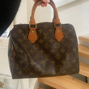 Speedy 25 Louis Vuitton - Säljer en klassisk brun handväska från Louis Vuitton med ikoniskt monogrammönster och detaljer i ljusbrunt skinn. Väskan har två handtag och dragkedja upptill. Perfekt för dig som gillar tidlös design och lyxiga accessoarer.. pris kan diskuteras 