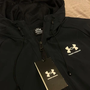 Svart vindjacka från Under Armour - Snygg svart vindjacka från Under Armour med huva och dragkedja framtill. Jackan har justerbara snören vid huvan och logga på bröstet. Perfekt för träning eller vardagsbruk när det blåser ute.
