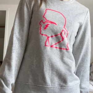 Grå sweatshirt från Karl Lagerfeld med rosa tryck - Säljer en grå sweatshirt från Karl Lagerfeld med ett stort rosa tryck av Karl Lagerfelds profil på bröstet. Tröjan har rund halsringning och långa ärmar. Perfekt för en cool och avslappnad stil.