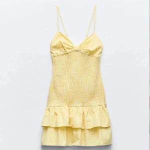 Gul kortklänning med volang och rosett - Supersöt gul kortklänning med smala axelband, volang nertill och rosett i ryggen. Klänningen har ett ljust rutigt mönster och är perfekt för varma dagar. Sitter figurnära upptill och har en luftig kjol.