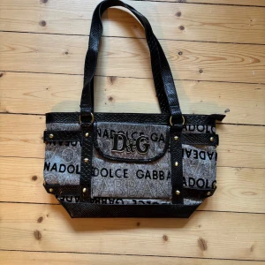 Svart och grå axelväska från Dolce & Gabbana - Säljer en snygg axelväska från Dolce & Gabbana i svart och grått med tydlig logga och textmönster. Väskan har svarta detaljer, dubbla axelband och dragkedja upptill. Perfekt för dig som vill ha en statement-väska med plats för allt du behöver.
