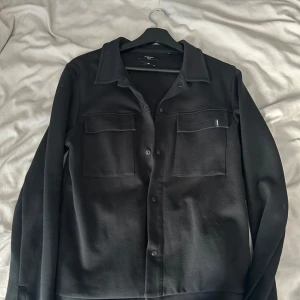 Svart tröja från Jack&jones - Säljer en svart tröja med klassisk krage och knäppning framtill. Tröjan  har två stora bröstfickor med lock och en stilren design. Perfekt för att addera en extra touch till din outfit.