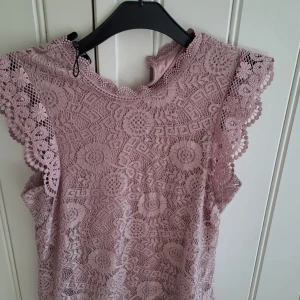 Ljuslila spetsblus från Pieces - Säljer en ljusrosalila blus från Pieces i vacker spets med blommigt mönster. Blusen är ärmlös med dekorativa volangdetaljer vid axlarna och har en rund halsringning. Perfekt för dig som gillar romantisk stil!