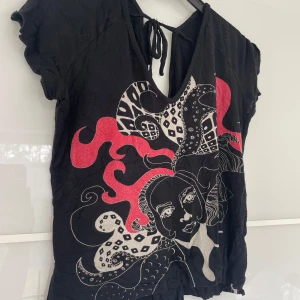 Svart t-shirt med konstnärligt tryck - Svart t-shirt med v-ringning och knytning i nacken. Framsidan har ett stort konstnärligt tryck i rött, vitt och grått med ansikte och abstrakta former