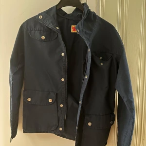 Mörkblå jacka från Fjällräven - Mörkblå vintage Fjällräven tonårsjacka från 80-talet med huva och flera fickor framtill. Jackan har både dragkedja och tryckknappar samt ett snyggt lädermärke på ärmen. Perfekt för dig som söker vintage/retro plagg.