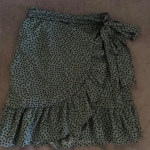 Grön stilren sommarkjol - Säljer en superfin grön omlottkjol med svarta prickar och volangkant. Kjolen knyts i midjan med en rosett. Perfekt för skolavslutning!🌞Passar XS/S 