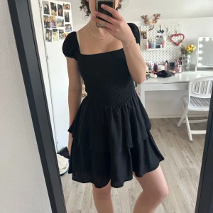 Svart volangklänning från Divided H&M - Supersöt svart kortklänning från Divided H&M med smockad överdel och puffärmar. Klänningen har volanger i kjolen och fyrkantig ringning. Perfekt för dig som gillar en feminin och trendig stil.