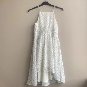 Vit blommig sommarklänning från H&M - Söt vit klänning från H&M med tunna axelband och små blå blommor. Klänningen har resår i midjan och är tillverkad i ett lätt, luftigt material. Perfekt för varma dagar och har en flowy passform med något längre bakstycke.