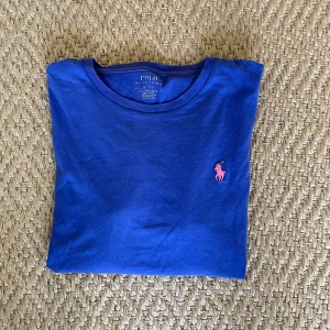 Blå t-shirt från Polo Ralph Lauren - Säljer en klassisk blå t-shirt från Polo Ralph Lauren med rosa broderad logga på bröstet. T-shirten har rund halsringning och är kortärmad. Perfekt för en stilren och avslappnad look.