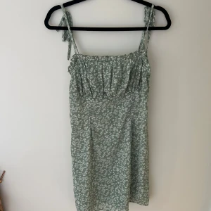 Grön blommig sommarklänning med knytband - Söt grön klänning med vita småblommor och smala axelband som knyts upptill. Klänningen har rynkad överdel och är i en lätt, luftig modell som passar perfekt för varma dagar.