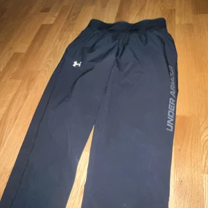 Svarta träningsbyxor från Under Armour - Svarta träningsbyxor från Under Armour med logga på låret och text längs benet. Byxorna har resår i midjan och dragkedjor vid bensluten för enkel av- och påtagning. Perfekta för sport eller gym.