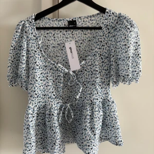 Blommig blus med knyt  - Supersöt blommig blus från Gina Tricot i vitt och blått. Blusen har korta puffärmar, knyt framtill och volang nertill. Perfekt för vår och sommar, med ett lätt och luftigt material. Storlek XS, oanvänd med prislapp kvar