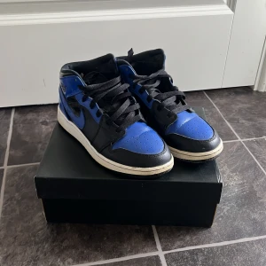 Nike Air Jordan 1 Mid svart/blå - Säljer ett par Nike Air Jordan 1 Mid i svart och blått med vita sulor. Klassisk high-top design med snörning och perforerad tå. Ikonisk Jumpman-logga på plösen och Swoosh på sidan. Perfekta för dig som gillar streetwear och sneakers. Skickas i orginalförpackningen. Använt skick men inget som syns om man inte kollar nära. Ny pris 2.600