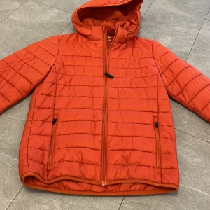 Orange pufferjacka från Everest - Säljer en orange pufferjacka från Everest med huva och dragkedja framtill. Jackan har två fickor med dragkedja och quiltad design. Perfekt för kyligare dagar. 