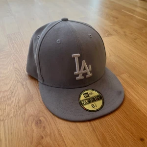 Grå LA keps från New Era - Säljer en grå keps från New Era med LA-logga broderad framtill. Modellen är 59FIFTY och har en klassisk, rak skärm. Kepsen är i bomull och passar perfekt för dig som gillar sportig stil.
