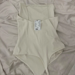 Beige bodysuit med tunna axelband - Säljer en stilren beige bodysuit med tunna axelband. Perfekt för lager-på-lager eller att bära som den är. Enkel och elegant design som passar till många olika outfits.