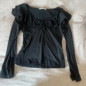 Svart långärmad topp med volang från Odd Molly - Supersnygg svart långärmad topp från Odd Molly med volang längs ringningen och knyt framtill. Perfekt för dig som gillar romantiska detaljer och en feminin stil.