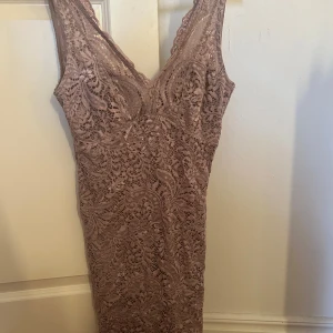 Ljusbrun spetsklänning med v-ringning - Säljer en elegant ljusbrun/ beige spetsklänning med djup v-ringning och figurnära passform. Klänningen har ärmlös design och vackert spetsmönster över hela plagget. Perfekt för speciella tillfällen!