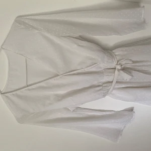 Vit klänning - Säljer en vit omlottklänning med knytband i midjan och diskret prickigt mönster. Klänningen har långa ärmar och volangdetaljer nertill