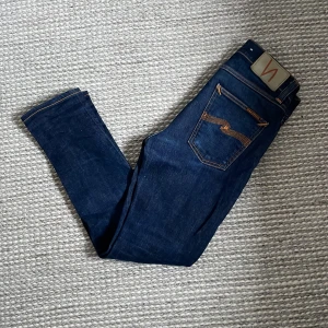 Nudie Jeans - Säljer ett par mörkblå jeans från Nudie Jeans med klassiska orange sömmar och femficksmodell. Jeansen har slim passform och normal midja, tillverkade i denim med snyggt läderpatch bak. Modell lean Dean sitter slim. W 27 L 28.