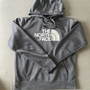 Grå hoodie från The North Face - Säljer en stilren grå hoodie från The North Face med stor vit logga på bröstet. Tröjan har luva med snöre och en stor magficka.