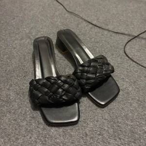 Svarta flätade sandaler med låg klack - Säljer ett par svarta sandaler med fyrkantig tå och låg klack. Ovandelen har en bred, flätad rem i syntetmaterial som ger en trendig look. Perfekta att styla till både jeans och klänning.
