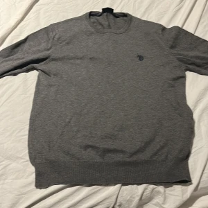 Grå tröja från U.S. Polo Assn. - Säljer en stilren grå tröja från U.S. Polo Assn. med rund halsringning och diskret logga broderad på bröstet. Tröjan har långa ärmar och ribbade muddar vid ärmslut och nederkant. Perfekt för en clean och enkel look.