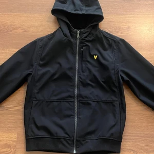 Svart vindjacka med huva från Lyle & Scott - Snygg svart vindjacka från Lyle & Scott med huva och dragkedja framtill. Jackan har två fickor och den klassiska gula loggan på bröstet. Perfekt för blåsiga dagar och enkel att matcha med det mesta.