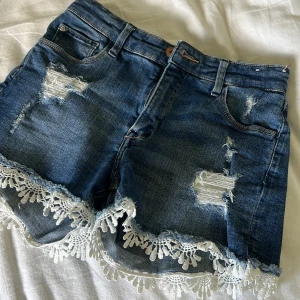 Jeansshorts med spetskant - Mörkblå jeansshorts med slitna detaljer och vit spetskant längst ner. Klassisk femficksmodell med bälteshällor och knappgylf. Perfekta för sommardagar när du vill ha något extra fint.