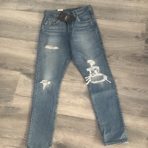Levis 501 blå jeans med slitningar - Klassiska blå Levis 501 jeans med raka ben och slitna detaljer på framsidan. Jeansen har hög midja och femficksmodell, tillverkade i robust denim. Perfekta för en avslappnad och trendig look.