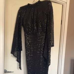 Svart glittrig långklänning med capeärmar - Säljer en svart långklänning med glittrigt mönster och höga capeärmar. Klänningen har en elegant och figurnära passform med hög hals och är perfekt för festliga tillfällen. Materialet är syntet och ger en lyxig känsla. Endast använd vid två tillfällen, passar en S/M i storlek. 