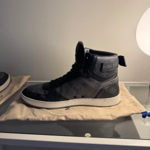 Svarta Louis Vuitton sneakers med monogram - Säljer ett par svarta high-top sneakers från Louis Vuitton med klassiskt monogrammönster. Skorna har snörning, vit platt sula och detaljer i skinn. Perfekta för dig som vill ha en lyxig och stilren look.