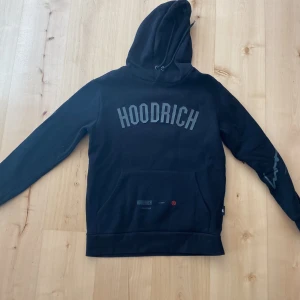 Hoodrich Hoodie - Säljer en svart hoodie från Hoodrich med stor logga framtill och textdetaljer på ryggen. Tröjan har huva, magficka och tryck på ena ärmen.