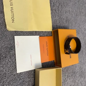 Svart armband från LV - Stilrent svart armband  med klassisk LV-logga i guld på framsidan. Armbandet är gjort i läder och har en enkel, elegant design. Perfekt accessoar för att lyfta din outfit.(F4KE och helt ny)