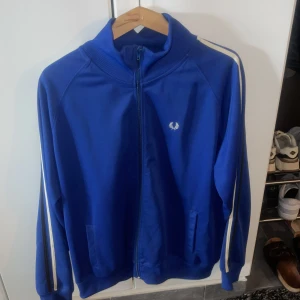 Blå track jacket från Fred Perry - Säljer en blå track jacket från Fred Perry med vita ränder längs ärmarna och dragkedja framtill. Jackan har ribbad krage, muddar och nederkant samt två fickor. Klassisk logga broderad på bröstet.