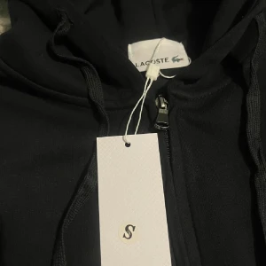 Svart hoodie från Lacoste med dragkedja - Svart hoodie från Lacoste med dragkedja framtill och huva med dragsko. Klassisk krokodillogga på bröstet och två fickor framtill. S 