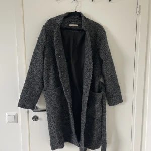 Grå/svart Kappa - Grå/svart aktig kappa från Gina Tricot i Wool Blend material. Skärpet sitter löst, men kan sys fast en ögla om man vill ha det.  Lite ”fnasig”, men ska också typ se ut så i modellen.