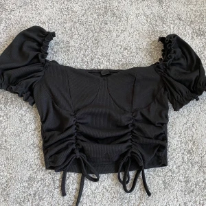 Svart topp med detaljer - En svart blus med detaljer som går att reglera. Är endast använd 1-2 gånger så är i nyskick. Säljs pga att den inte kommer till användning. Nypris runt 130kr & säljs för 70kr, priset kan diskuteras. Köparen står för frakten! 