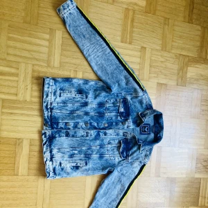 Jeans jacka  - Jeansjacka i storlek L med svarta och gula stripes längs armarna. Nyskick.  Modellen på bilden är 185 cm lång. Vikt 88 kg  