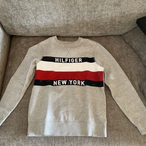 Sweatshirt  - Säljer denna grå sweatshirt från Tommy Hilfiger då jag växt ur den. Plagget är i mycket bra skick förutom lite färg förlust på det blåa på framsidan. Säljer till mycket bra pris.