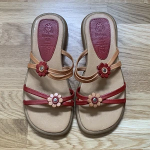 Somriga slip-ins - urgulliga och somriga slip-ins med blomdetaljer! Jättesköna och passar perfekt till sommarens alla outfits!🌺🌼🌸 Tillverkade av äkta skinn. Beige/brun med röda detaljer. Använda ett fåtal gånger! 