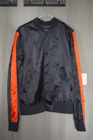 Svart bomberjacka med röda detaljer - Säljer denna svarta bomberjacka i glansigt material. Den har röda detaljer längs armarna. Den har bara blivit använd ett fåtal gånger och är så god som ny.