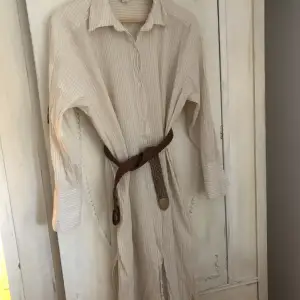 Vit och beige randig skjortklänning från H&M. Lång. Slitsar på sidan. Snyggt med bälte i midjan. Luftig i skön nu i sommar. Strl M