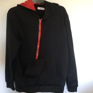 Rödsvart osymmetriskt hoodie stl M - Denna hoodie är svart med en röd dragkedja och är röd på insidan av luvan. Vid öppning av dragkedjan är ordet Swag i röda bokstäver. Tröjan är aldrig använd och i nytt skick. Den är i storlek M. 